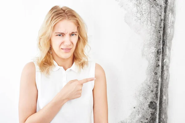 Wall fungus. Aspergillus. A beautiful girl in a white T-shirt points a finger at the black mold on the wall