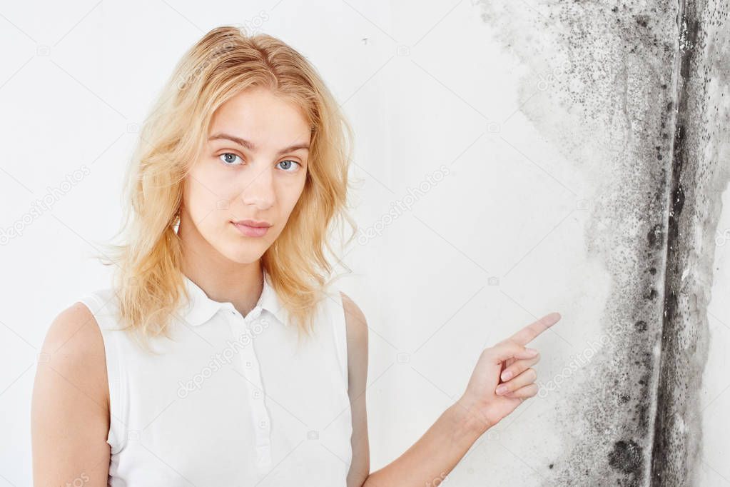 Wall fungus. Aspergillus. A beautiful girl in a white T-shirt points a finger at the black mold on the wall