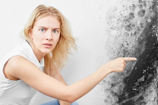 Wall fungus. Aspergillus. A beautiful girl in a white T-shirt points a finger at the black mold on the wall