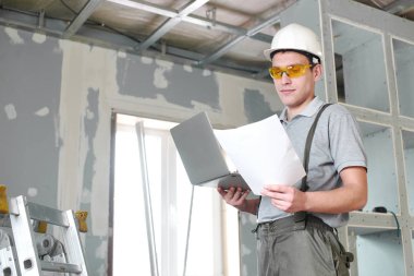 Oda onarım. Genç Oluşturucusu'nda kask ve gözlük ile bir dizüstü bilgisayar ve çizimler inşaat zemin üzerine onun elinde duruyor