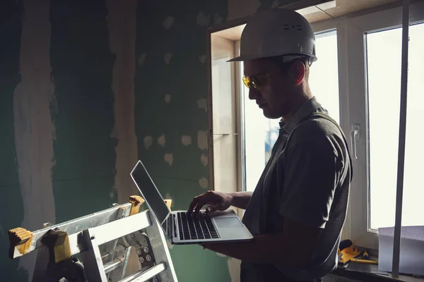 房间维修 戴头盔和眼镜的建筑商站在建筑工地的背景上 手里拿着一台笔记本电脑 — 图库照片