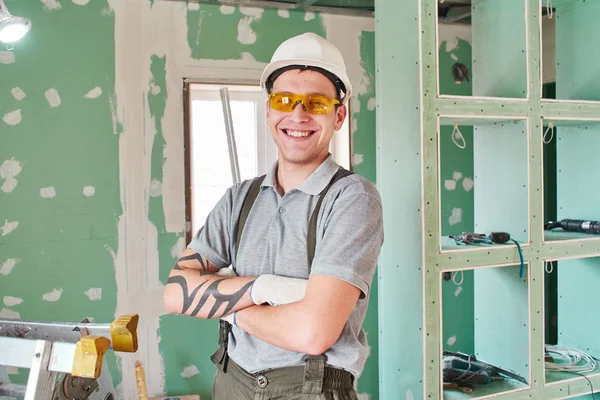 部屋を修復します ヘルメットと建設現場の背景に手を組んで立っているオーバー オールの若い笑顔ビルダー — ストック写真