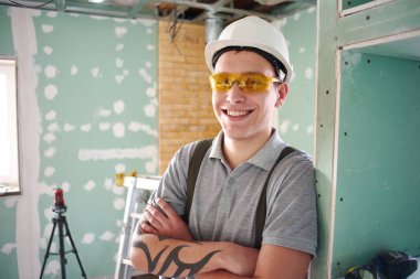 Oda onarım. Genç gülümseyen Oluşturucusu'nda bir kask ve inşaat alanında arka plan üzerinde katlanmış ellerle ayakta tulum