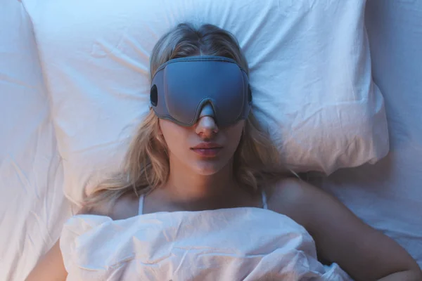 Sleeping girl in a sleep mask on an orthopedic pillow under night lighting, white bedclothes, top view