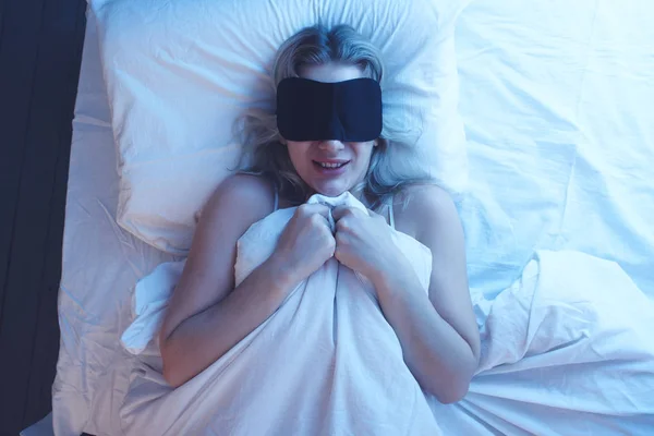 寝具を白い恐怖と夜の照明の下で整形外科枕を彼女の顔に恐怖の表情で睡眠マスクで女の子を寝て — ストック写真