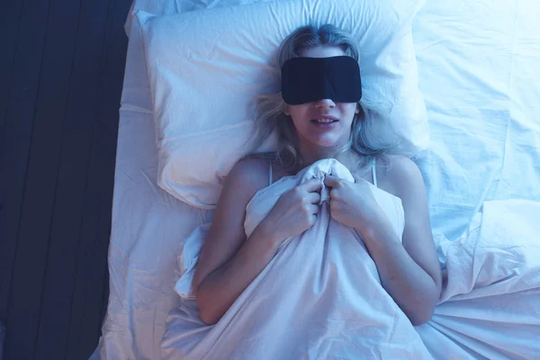 Nightmares. Sleeping girl in a sleep mask with an expression of fear and horror on her face on an orthopedic pillow under night lighting, white bedding.