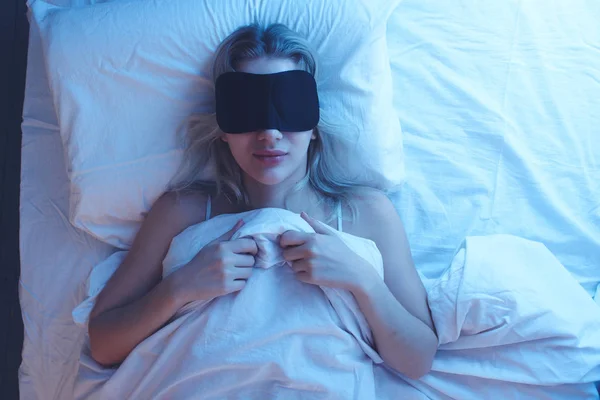 Nightmares. Sleeping girl in a sleep mask with an expression of fear and horror on her face on an orthopedic pillow under night lighting, white bedding.