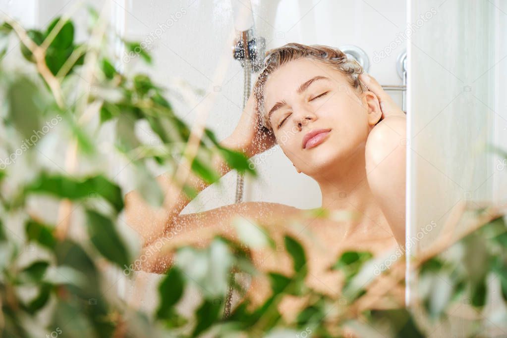 Beautiful satisfied European woman washes away shampoo from the head hair in bathroom, takes a shower and enjoys, smiling