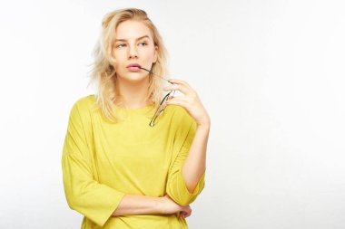 Pensive European stylish young woman in yellow clothes holding glasses near mouth, doubts what to choose, thinks, decides what to buy on white background clipart
