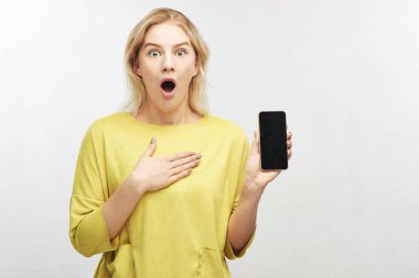 Cep telefonu ile şok Avrupa sarışın kadın elinde, çene düştü sürpriz, ekranda, gördükleri gelen duygular içinde şişkin gözler koruyamaz. Güzel kız beyaz studio izole portresi