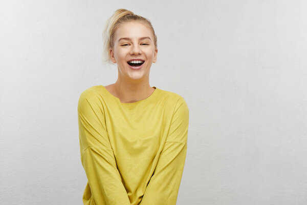 Joyful satisfied adult girl with funny ponytail hair wearing yellow casual clothes, laughing at joke in white studio, positive mood before meeting with friends