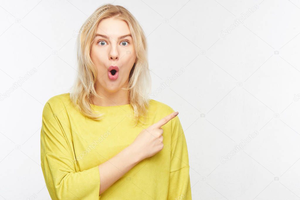 Surprised blond woman with open mouth points to upper right corner index finger, stunned advertise your product in yellow casual clothes on white background. Oh my god concept