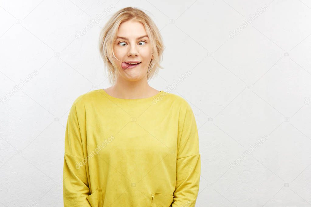A crazy blond girl in yellow clothes on white background squints eyes, sticks out tongue, makes funny fool face, does not want to grow up and be responsible