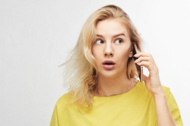 Portrait of shocked girl with open mouth talking mobile phone on white background with free advertising space. Woman in yellow casual outfit hears something amazing, she confused clipart