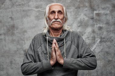 The pacified gray-haired grandfather folded his palms in prayer position, meditation, relaxation, forgiveness, keep calm. Gray studio background clipart