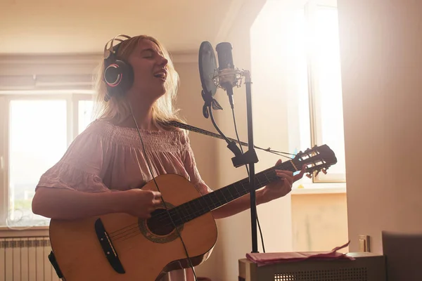 Beautiful blonde singer girl in headphones with a guitar in home recording studio sings a song into a microphone