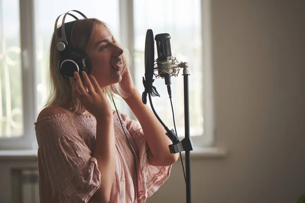 Beautiful blonde singer girl in headphones in home recording studio sings a song into a microphone, laughs and smiles close-up