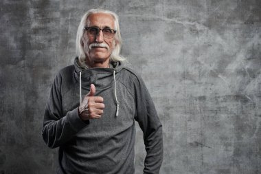 Bir bıyık ile modern gri saçlı Kafkas yaşlı adam başparmak yukarı ve gülümsüyor, stüdyoda gri duvar karşı neşeli mutlu yüz gösterir