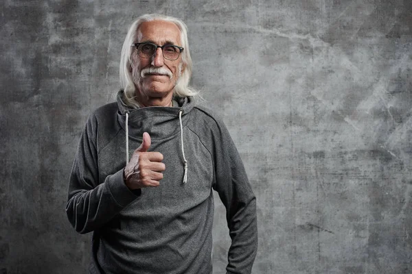 Bir bıyık ile modern gri saçlı Kafkas yaşlı adam başparmak yukarı ve gülümsüyor, stüdyoda gri duvar karşı neşeli mutlu yüz gösterir