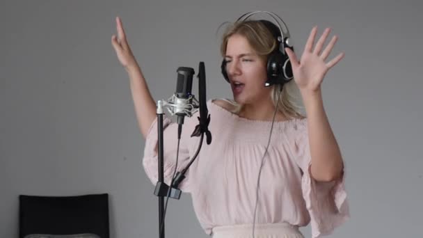 Hermosa cantante rubia en auriculares en el estudio de grabación en casa canta una canción en un micrófono, jugando, posando y bailando con una cara emocional expresiva — Vídeo de stock