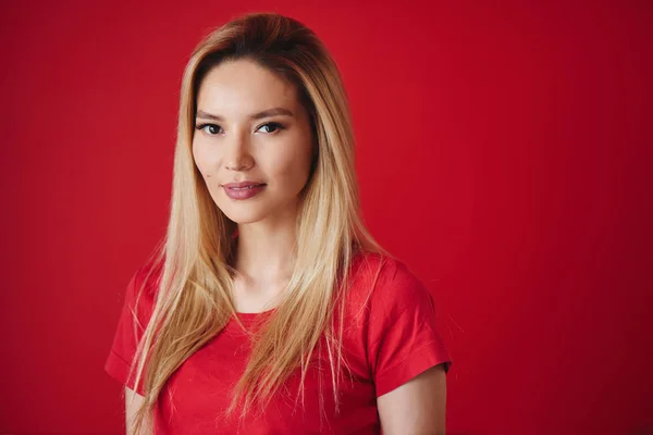Portrait of beautiful asian young woman in t-shirt. Attractive kazakh blonde girl smiling on red background in studio isolated