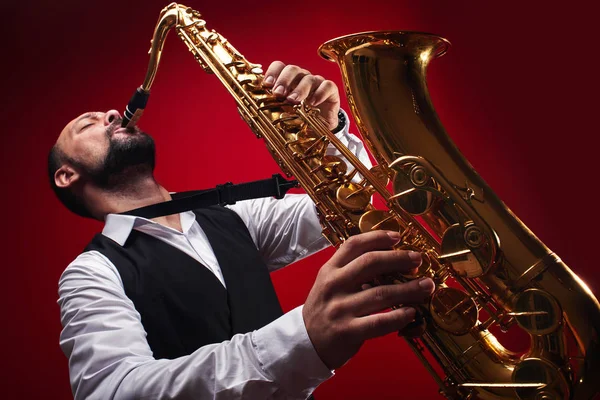 Portrait of professional musician saxophonist man in  suit plays jazz music on saxophone, red background in a photo studio, bottom view