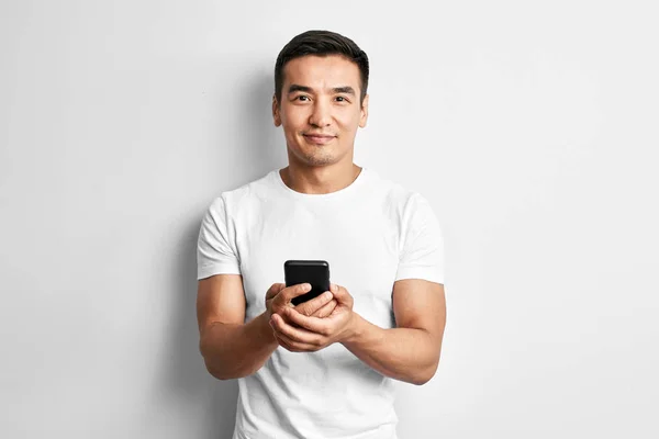 Beyaz stüdyoyakışıklı Asyalı adam sevinçle cep telefonu ekranına bakar gülümsüyor. Mutlu memnun adam rahat giyinmiş yazar ve akıllı telefonda sms okur