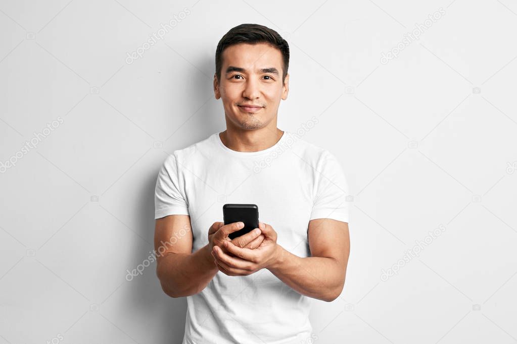 Handsome Asian guy in white studio smiles joyfully looks at the screen of mobile phone. Happy contented man dressed casual writes and reads sms on smartphone