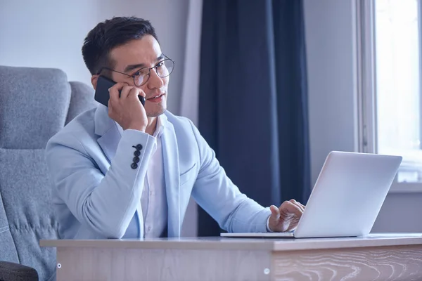 Asian Kazakh businessman in a suit and glasses with a laptop computer in the office speaks by mobile phone, solves business issues, negotiates a deal.