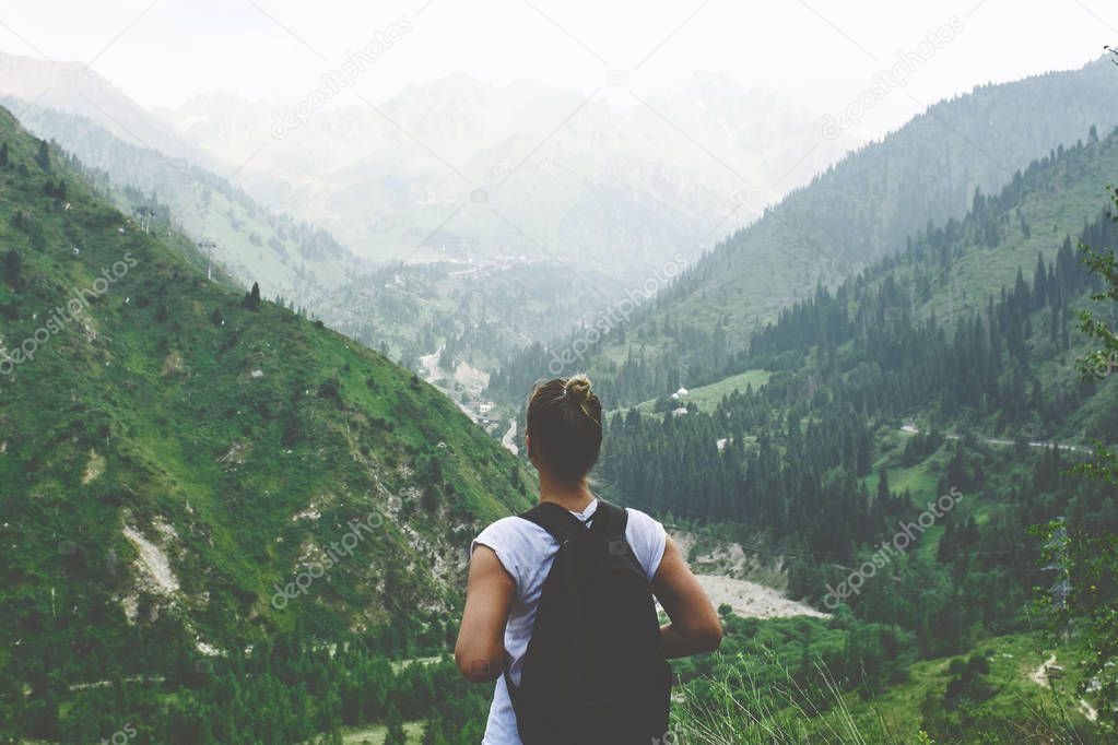 Young woman traveler with a backpack behind her back in Zailiysky Alatau in Almaty looks at the mountain landscape of summer Kazakhstan