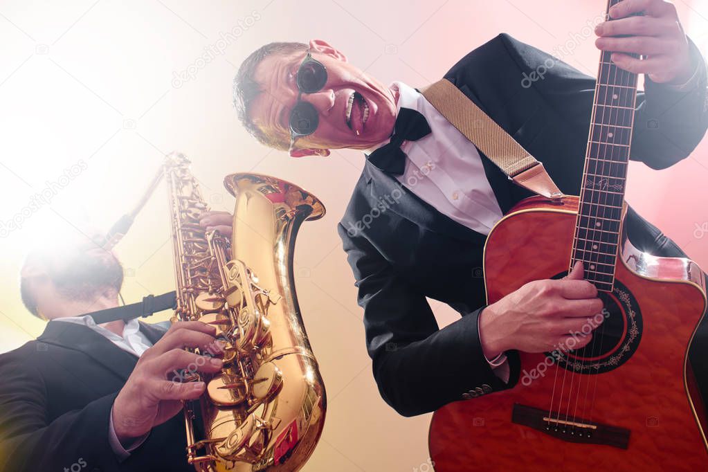 Group of two musicians, male jazz band, guitarist and saxophonist in classical costumes improvise on musical instruments in a studio stage lighting