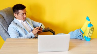 Komik çorap ve gözlük ile takım elbise yakışıklı Asyalı işadamı masaya ayaklarını koymak, ofiste çalışıyor, başarılı Kazak yöneticisi cep telefonu ile iş sorunları çözer