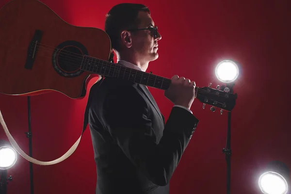 Portrait of classical musician with guitar in red studio with stage lighting. Guitarist in black glasses and suit with a bow tie improvises on instrument