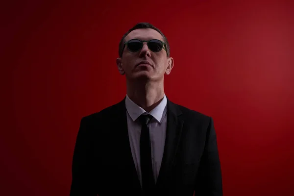 Portrait of a strict serious adult man in a suit and black glasses on a red studio background. Face control, night club security