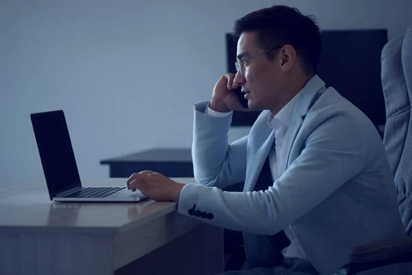 Asian Kazakh businessman in a suit and glasses with a laptop computer in the office speaks by mobile phone, solves business issues, negotiates a deal.