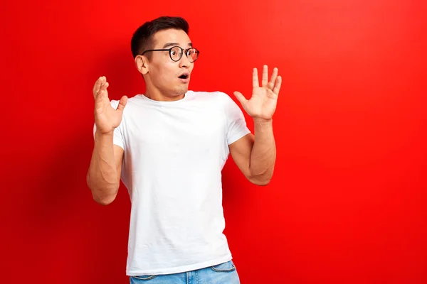 白いカジュアルな服を着たショックを受けたアジア人男性は 口を開けて手を上げて横を見る 赤いスタジオの背景に驚いた若いカザフスタンの学生の男 — ストック写真