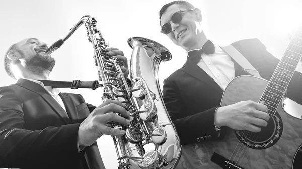 Group of two musicians, male jazz band, guitarist and saxophonist in classical costumes improvise on musical instruments in a studio black and white shot