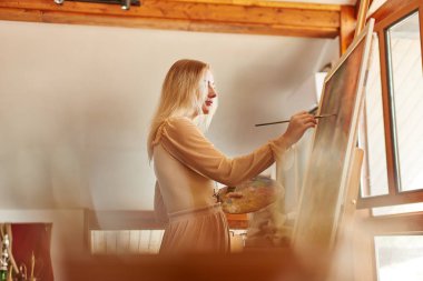 Güzel yetenekli genç kadın sanatçı şık yaratıcı sanat stüdyosu elinde palet ile bir şelon üzerinde fırça ve yağ ile bir resim boyar