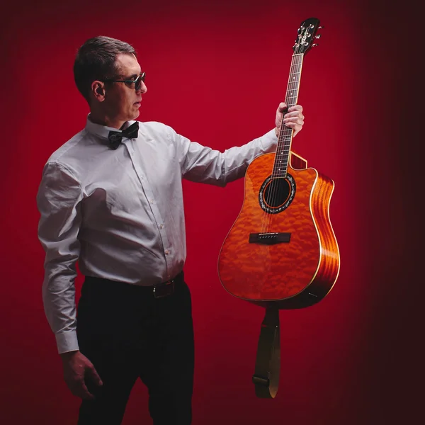 Portrait of classical musician with guitar in red studio. Guitarist in black glasses and a white shirt with a bow tie holds instrument in hand