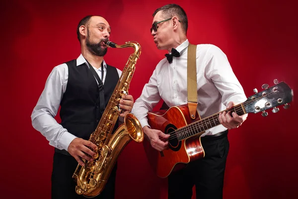 Group of two musicians, male jazz band, guitarist and saxophonist in classical costumes improvise on musical instruments in a studio on  red background