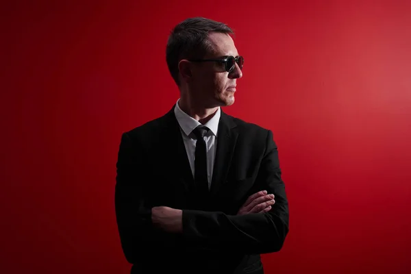Portrait of strict serious adult man in suit and black glasses looks away with crossed arms on red studio background. Face control, night club security