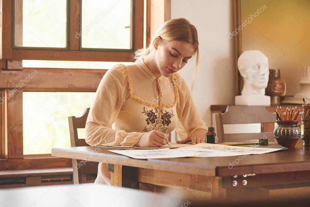 Creative young beautiful woman artist or poet sits at wooden table in art studio and writes  picture or letter in ink. Romantic stylish photo in warm beige colors