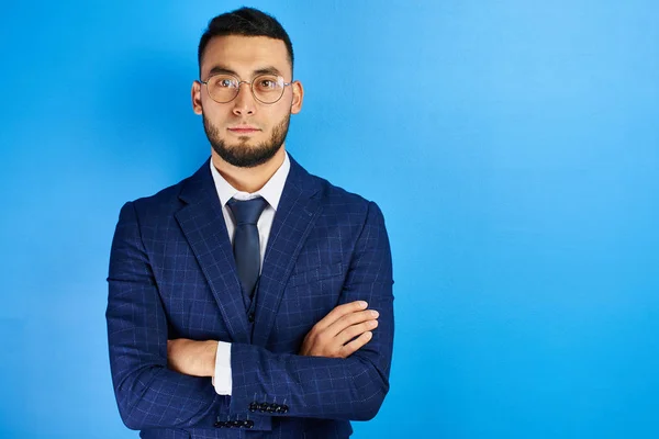 Portrait of serious handsome successful kazakh asian businessman in suit and glasses with arms crossed on blue studio background , isolated with copy space