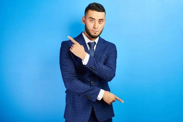 Asian Kazakh man in a business suit with a serious face shows fingers to the sides on copy space on a blue wall isolated