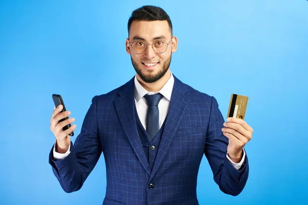 Asian Kazakh business bank representative man in suit and glasses smiles joyfully and holds mobile phone and credit card in hands on blue background isolated