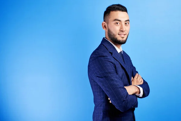Portrait of smiling handsome successful kazakh asian businessman in suit with arms crossed on blue studio background , isolated with copy space