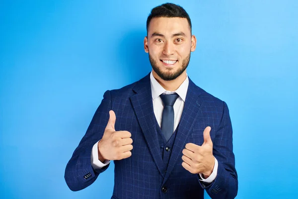 Successful Asian Kazakh businessman shows thumbs up and toothy smiles on a blue studio background. Isolated
