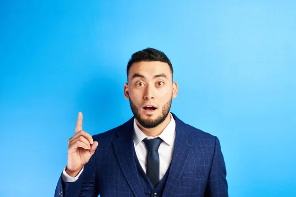 喜んだアジアのカザフスタンのビジネスマンは スーツで開いた口で人差し指を上げ 青いスタジオで孤立したネクタイを持っています — ストック写真