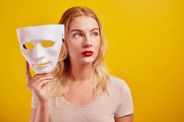 Emotional female person with white mask on yellow background. Internet fraud concept, anonymous, incognito, bipolar personality disorder, hypocris