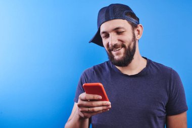 Şapkalı yetişkin beyaz sakallı adam cep telefonuyla oyun oynuyor ve mavi stüdyo arka planındaki bir çocuk gibi seviniyor, çocukça bir kavram.